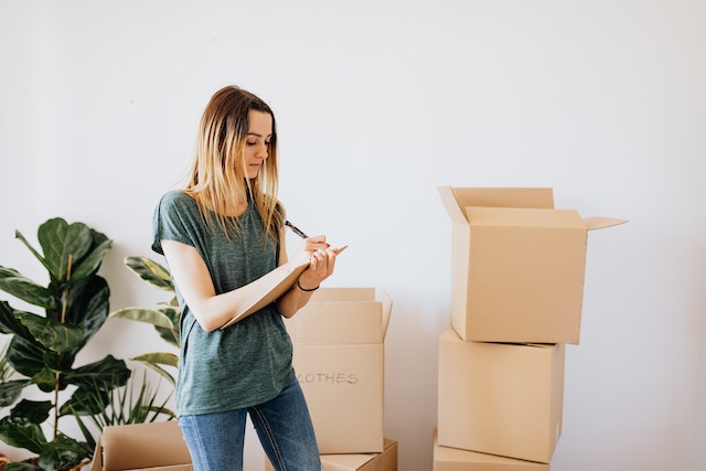 Person writing a list on a clipboard next to a stack of cardboard moving boxes